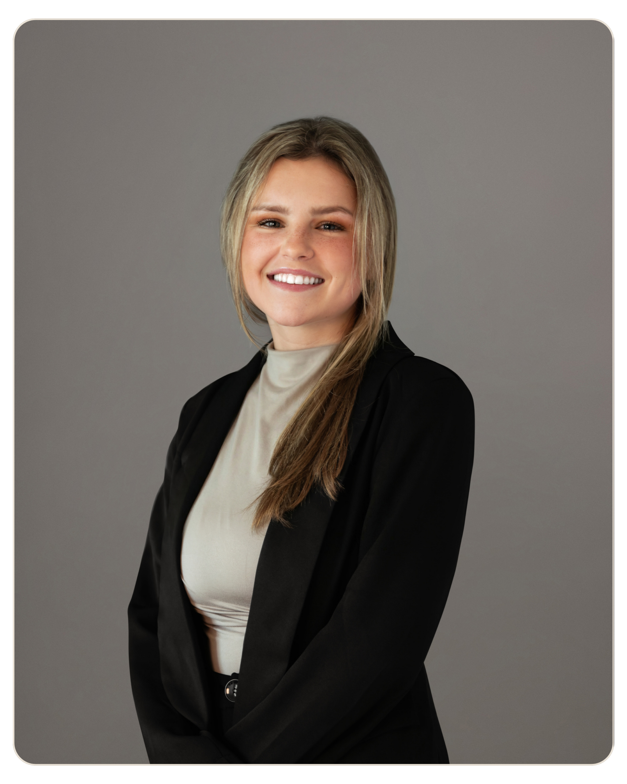 Smiling woman with blonde hair wearing a beige top and black blazer against a gray studio background.