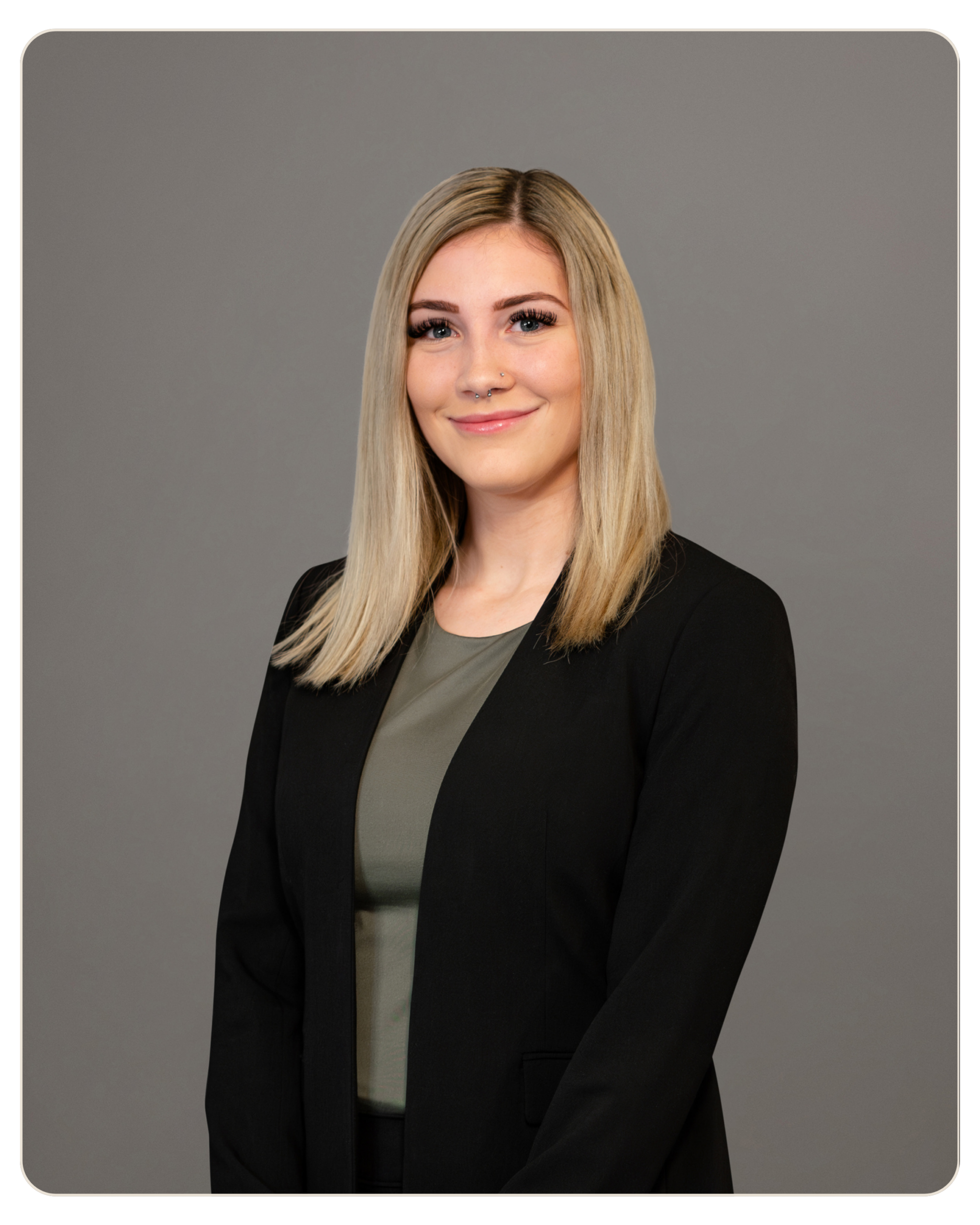 Nicole Laska Trost Professional headshot of a young woman with long blonde hair, wearing a black blazer over a gray-green top, smiling against a gray studio backdrop
